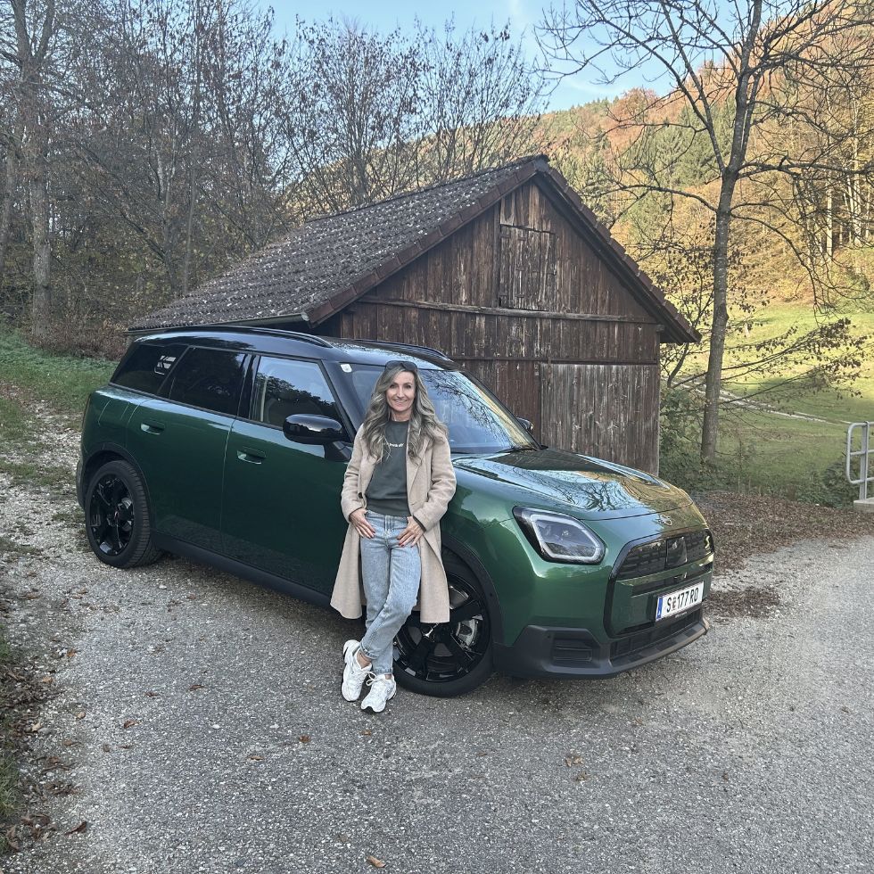 Grüner Mini Countryman und eine Frau vor einer Holzhütte.