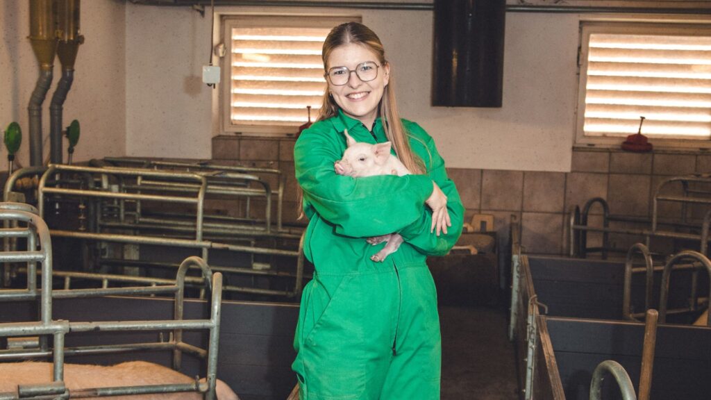 Junge Fraus steht in einem Stall mit einem Ferkel auf dem Arm.