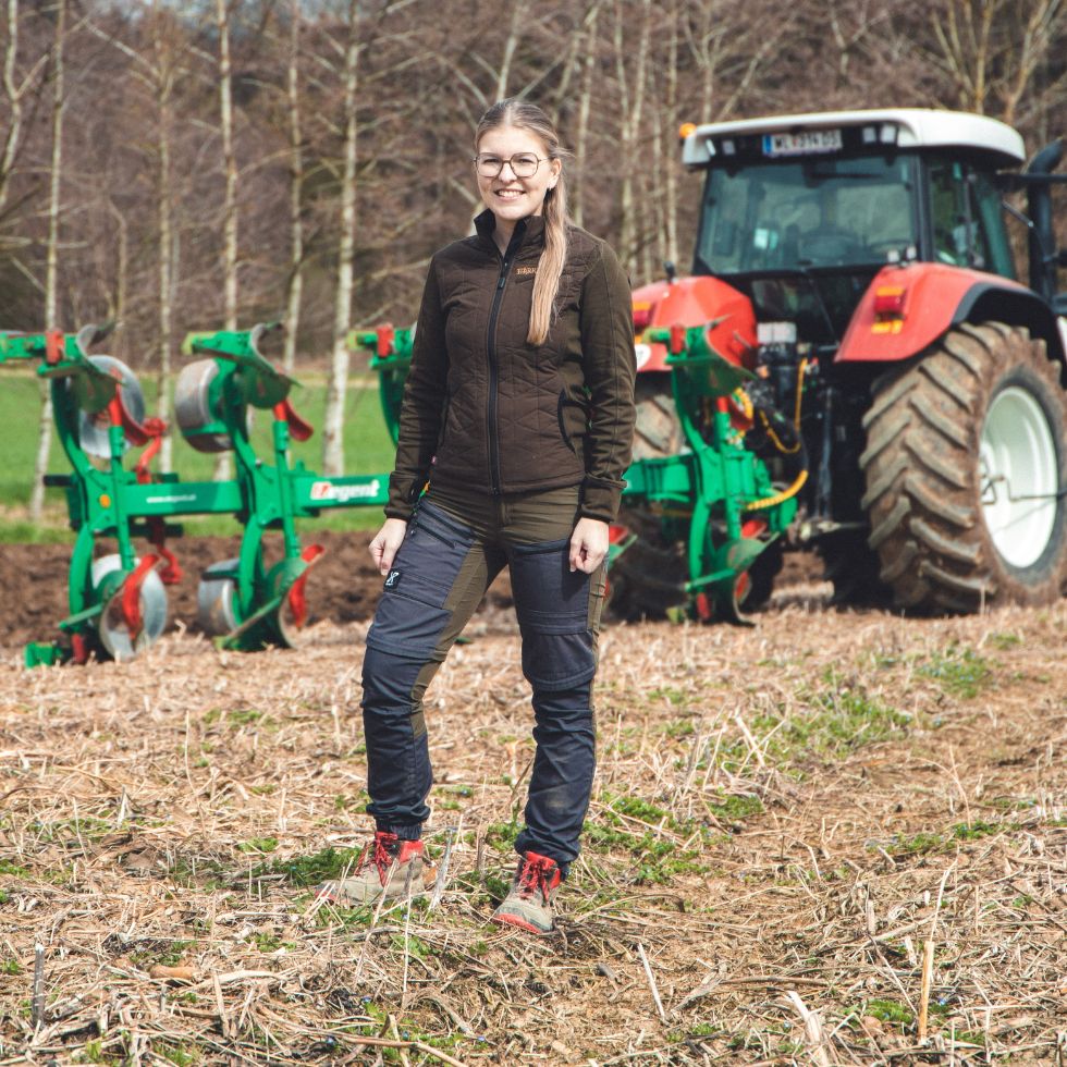 Junge Frau steht auf dem Feld mit einem Traktor im Hintergrund.