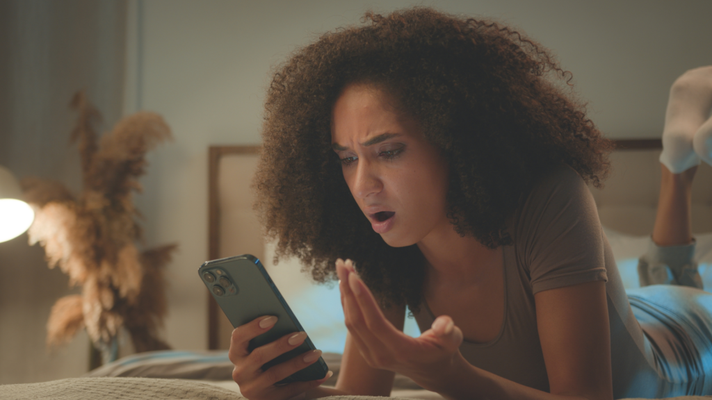 Junge Frau liegt auf dem Bett und schaut angewidert auf ihr Smartphone.
